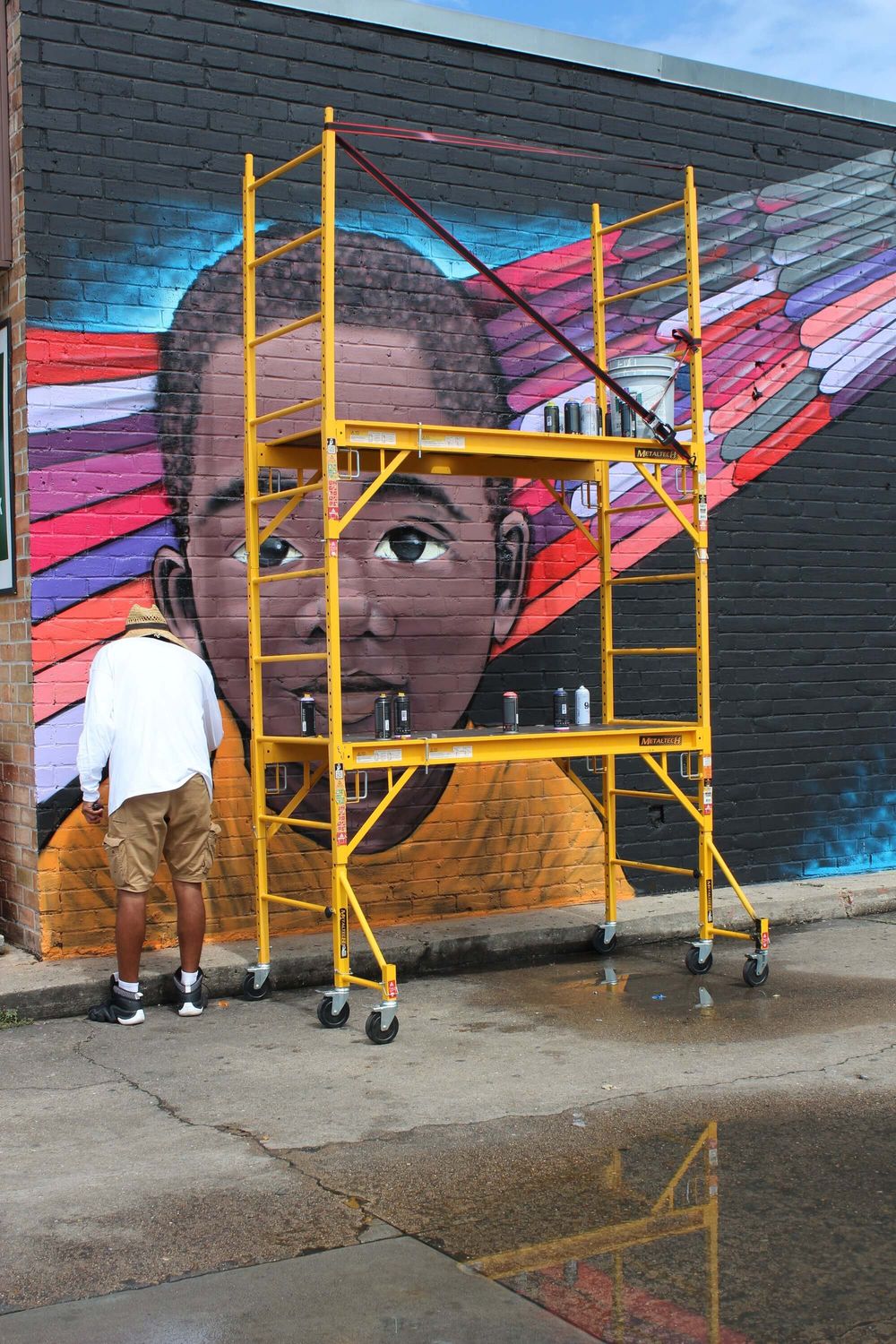 An artist works from scaffolding on a large portrait mural on a brick wall