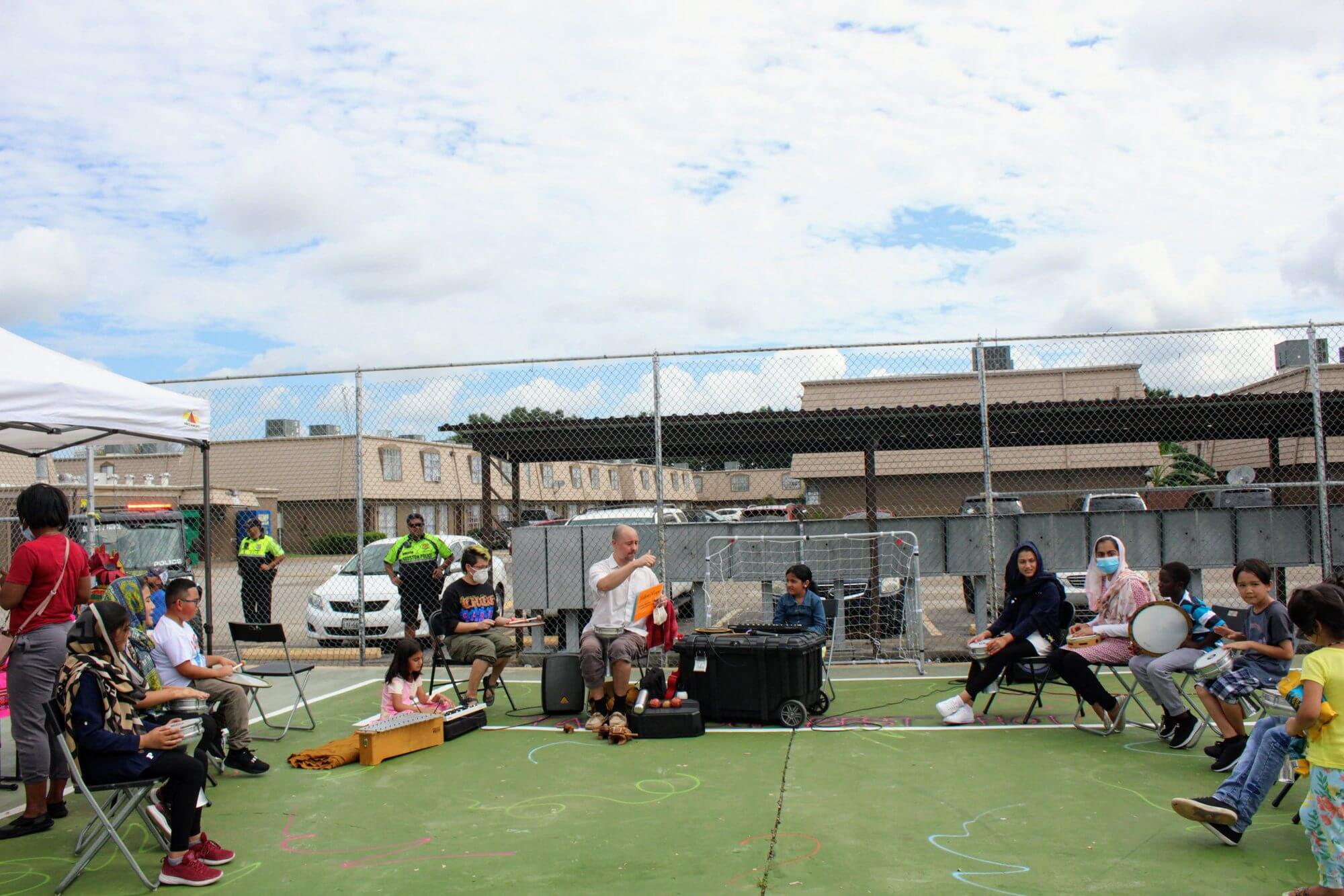 Neighbors of all ages seated in a circle with drums and percussion instruments at an outdoor community session