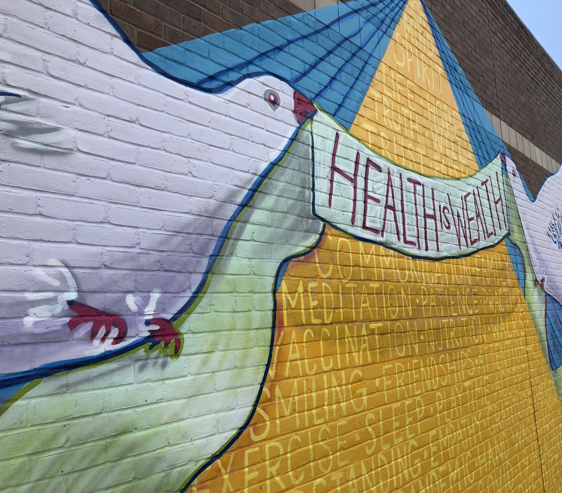 Close view of a mural of doves carrying a Health is Wealth banner over a list of wellbeing words