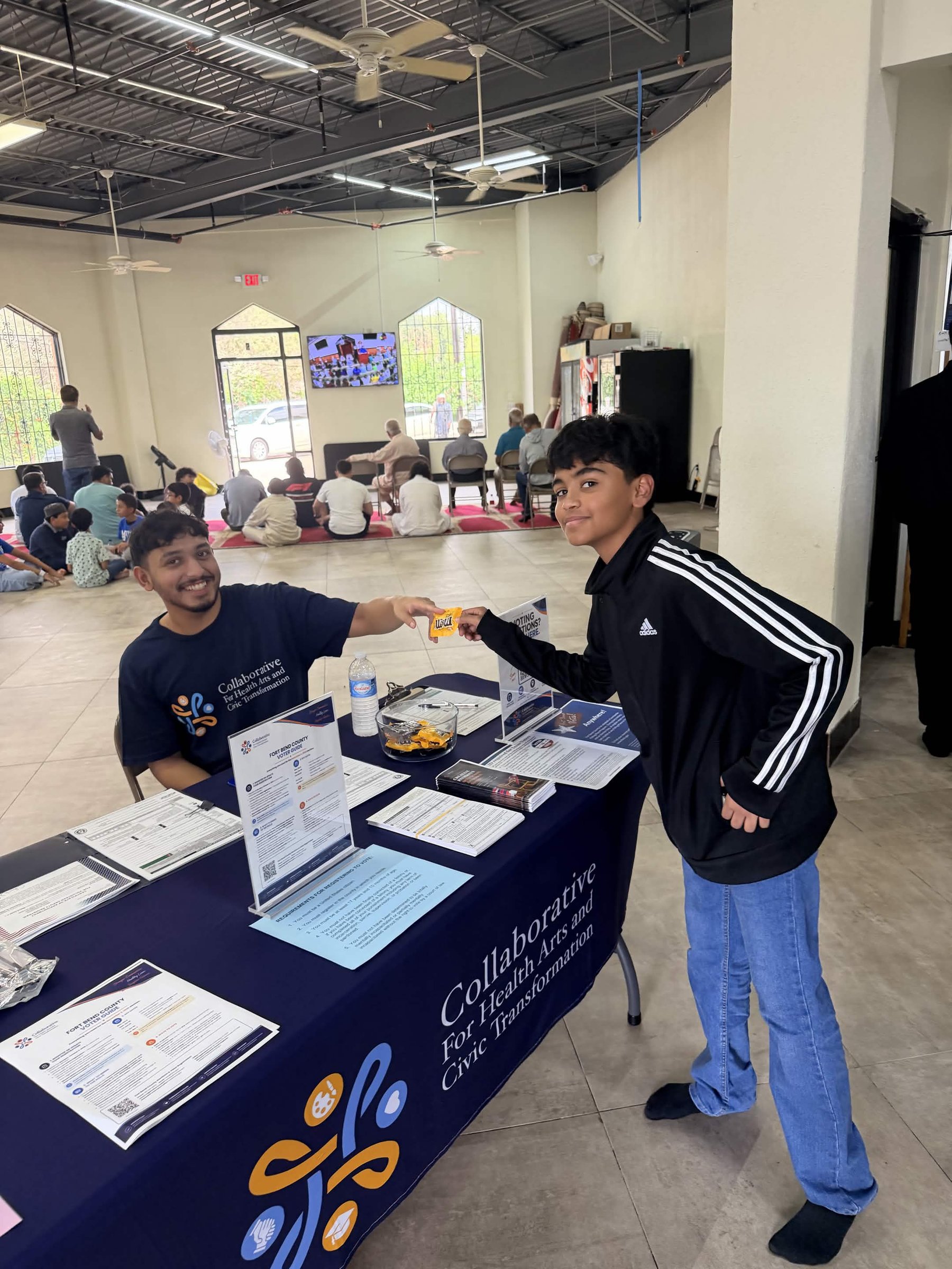 A volunteer at the Collaborative table with a visitor, voter guides and registration forms laid out